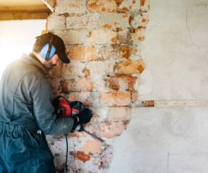 Mature worker demolishing wall with drill at house